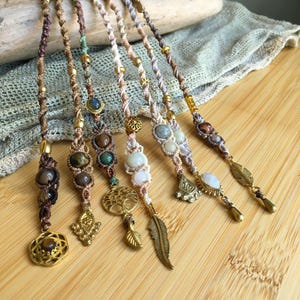 May include: Six macrame bracelets featuring braided cord in shades of brown, beige, and cream. Each bracelet is adorned with various round gemstones, gold-toned metal beads, and charms including a tree of life, a feather, and geometric shapes.