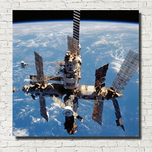 以下が含まれることがあります： 地球上空の軌道にある宇宙ステーションの正方形のプリント。ステーションには複数のソーラーパネルアレイとモジュールがあります。下の地球は、雲の形成を伴う青と白です。プリントは白いレンガの壁にあります。