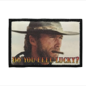 May include: A black and white embroidered patch featuring a man wearing a cowboy hat and a mustache. The man is holding a cigar in his mouth and the text "Do you feel lucky?" is printed below him.