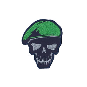 May include: A black and green embroidered patch of a skull wearing a green beret.