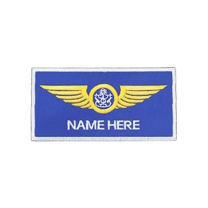 May include: A blue rectangular patch with a white border. The patch features a gold emblem of wings and a central design, with the text "NAME HERE" in white.