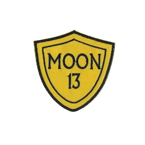 May include: Yellow embroidered patch in the shape of a shield with a black outline. The patch says "MOON 13" in black lettering.