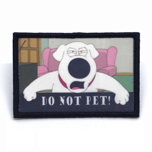 May include: A white dog with a black nose and a pink collar is shown in a close-up image. The dog is looking at the camera with a serious expression. The dog is sitting in a chair and has a sign in front of it that says "DO NOT PET!"