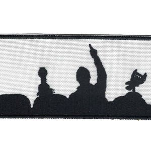 May include: Black and white embroidered patch showing a silhouette of three figures in front of a screen. The figures are standing and appear to be performing on stage. The patch is rectangular and has a white background with black stitching.