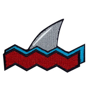 May include: A red and blue embroidered patch with a gray shark fin sticking out of the water. The water is represented by a blue and white zig-zag pattern.