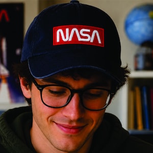 Pu&ograve; includere: Cappellino da baseball blu navy con il logo NASA in lettere bianche su un rettangolo rosso. Il cappellino &egrave; indossato da una persona con occhiali con montatura nera. Un poster di uno space shuttle &egrave; visibile sullo sfondo.