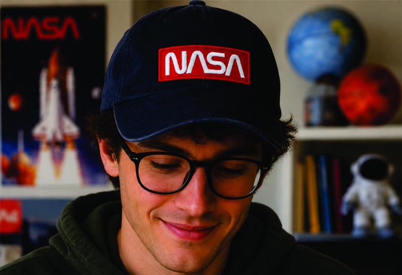 Pu&ograve; includere: Cappellino da baseball blu navy con il logo NASA in lettere bianche su un rettangolo rosso. Il cappellino &egrave; indossato da una persona con occhiali con montatura nera. Un poster di uno space shuttle &egrave; visibile sullo sfondo.