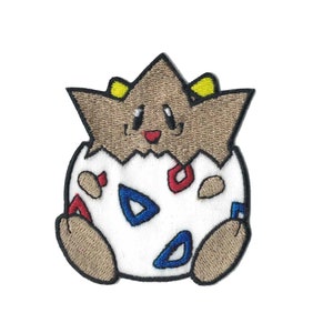 May include: An embroidered patch featuring a brown and yellow Pokemon character, known as a "Togepi", hatching from a white egg with red and blue markings.