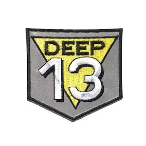 May include: A grey and yellow embroidered patch with the text "DEEP 13" in a triangular shape.