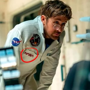 Pu&ograve; includere: Una tuta spaziale NASA di colore chiaro con il logo e le toppe della NASA. La tuta spaziale ha una tasca con il logo NASA in rosso. L'astronauta indossa gli occhiali e guarda qualcosa.