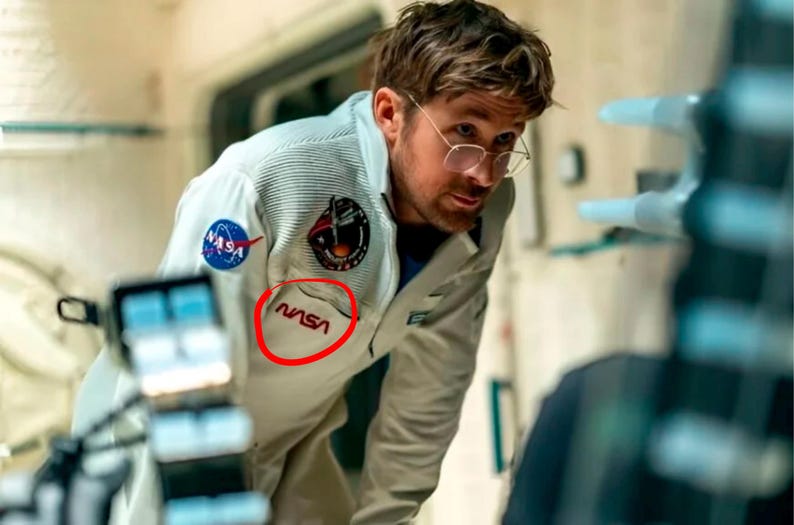 Pu&ograve; includere: Una tuta spaziale NASA di colore chiaro con il logo e le toppe della NASA. La tuta spaziale ha una tasca con il logo NASA in rosso. L'astronauta indossa gli occhiali e guarda qualcosa.