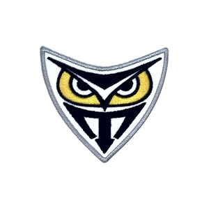 May include: A black and white embroidered patch with a stylized owl face. The owl has yellow eyes and a white beak. The patch is shaped like a triangle with a gray border.