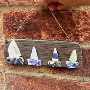May include: A driftwood sign with four ceramic sailboat decorations. Each boat has a white sail and a base with blue and white patterns. The sign is hung with twine, ideal for coastal-themed interiors.