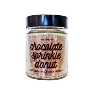 Może przedstawiać: Szklana świeca w słoiku z czarną pokrywką. Świeca jest oznaczona jako "chocolate sprinkle donut" i "nook + burrow". Świeca jest jasnobrązowa i ma białą etykietę z brązowym tekstem.