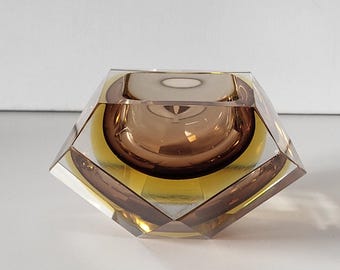 May include: A geometric glass ashtray with a brown and yellow color scheme. The ashtray has a faceted design and a square shape.