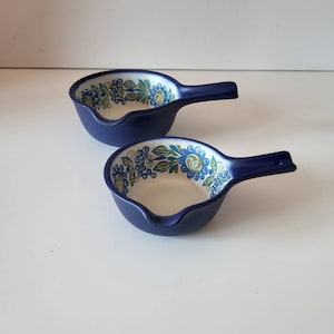 Puede incluir: Dos cuencos de cerámica azul marino con asas. El interior de cada cuenco presenta un estampado floral en tonos azules, verdes y blancos. Los cuencos están sobre un fondo blanco.