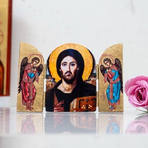 May include: A religious triptych featuring a central image of Jesus with a halo, flanked by angels in red and blue robes. The artwork is set against a gold background. Pink and white roses are in the foreground.