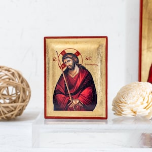 Può includere: Un'icona religiosa raffigurante una figura in abito rosso, con un bastone e un'aureola. L'icona ha uno sfondo dorato e una cornice rossa. Sono presenti anche una sfera decorativa e un oggetto floreale bianco.