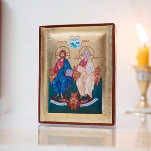 May include: A religious icon featuring two figures in robes, one in blue and the other in white, with gold leaf accents. The icon has text in gold lettering and is set against a white background. A lit candle and a ball of twine are in the background.