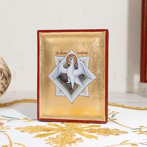 May include: A religious icon featuring a white dove within a starburst design, set against a gold background. The icon has a red border and Greek text at the top. The image is on a white surface with gold embroidery.