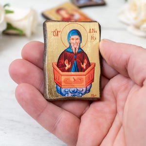 May include: A small, rectangular religious icon featuring a figure with a halo, wearing a blue head covering and red robe. The icon is set against a gold background and framed with dark wood. The image is held in a hand.