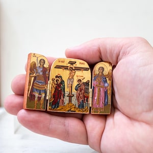 Może przedstawiać: Mały, zawiasowy drewniany tryptyk przedstawiający ikonografię religijną. Centralny panel przedstawia ukrzyżowanie, flankowane panelami z postaciami anioł&oacute;w. Dzieło sztuki jest szczeg&oacute;łowe z żywymi kolorami i złotymi akcentami, trzymane w dłoni.