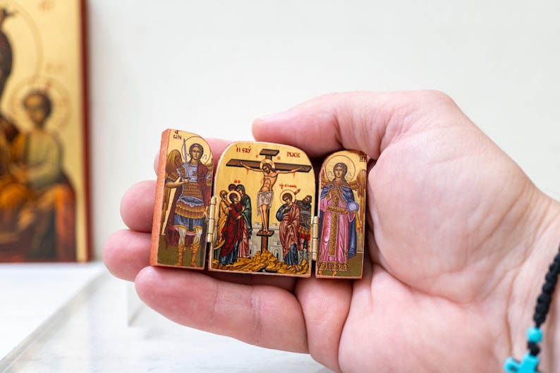 Może przedstawiać: Mały, zawiasowy drewniany tryptyk przedstawiający ikonografię religijną. Centralny panel przedstawia ukrzyżowanie, flankowane panelami z postaciami anioł&oacute;w. Dzieło sztuki jest szczeg&oacute;łowe z żywymi kolorami i złotymi akcentami, trzymane w dłoni.