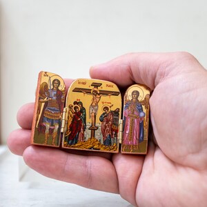 Może przedstawiać: Mały, zawiasowy drewniany tryptyk z ikonografią religijną. Dzieło przedstawia postacie z aureolami, aniołami i scenami ukrzyżowania. Panele są pomalowane żywymi kolorami i złotymi akcentami, prezentując szczeg&oacute;łowy, tradycyjny styl.