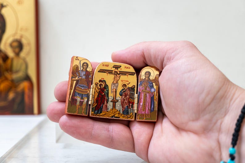 Może przedstawiać: Mały, zawiasowy drewniany tryptyk z ikonografią religijną. Dzieło przedstawia postacie z aureolami, aniołami i scenami ukrzyżowania. Panele są pomalowane żywymi kolorami i złotymi akcentami, prezentując szczeg&oacute;łowy, tradycyjny styl.