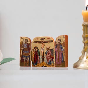Może przedstawiać: Ikona religijna przedstawiająca scenę ukrzyżowania, otoczona wizerunkami anioł&oacute;w. Ikona wykonana jest z drewna i posiada szczeg&oacute;łowe zdobienia ze złotymi akcentami. Po prawej stronie zapalona świeca w złotym świeczniku.