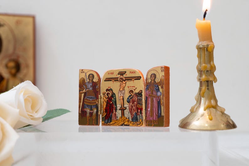 Może przedstawiać: Ikona religijna przedstawiająca scenę ukrzyżowania, otoczona wizerunkami anioł&oacute;w. Ikona wykonana jest z drewna i posiada szczeg&oacute;łowe zdobienia ze złotymi akcentami. Po prawej stronie zapalona świeca w złotym świeczniku.