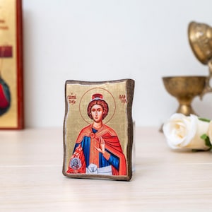 May include: A religious icon featuring a saint with a halo, wearing a red cloak and blue garments. The icon has gold accents and is set against a gold background. The icon is on a wooden base. Other religious items are in the background.
