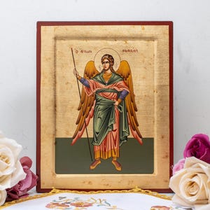 Peut inclure: Icône religieuse encadrée représentant un ange avec des ailes dorées tenant une lance. L'ange porte une robe verte et rose. L'icône a des accents de feuilles d'or et est posée sur un fond blanc. Des roses et un tissu décoratif sont au premier plan.