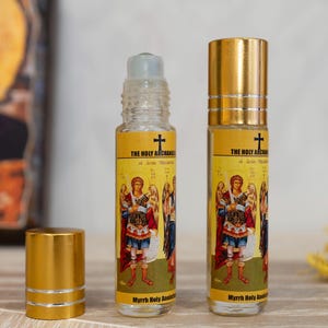 Op de afbeelding: Twee doorzichtige glazen flessen met gouden deksels en etiketten. De etiketten tonen een religieus icoon van een heilige aartsengel op een gouden achtergrond met de tekst "The Holy Archangel" en "Myrrh Holy Anointing Oil".