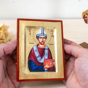 Op de afbeelding: Een rechthoekig religieus icoon met een geschilderde figuur van een heilige in een blauw en rood gewaad met een gouden achtergrond, omlijst door een rode rand. De heilige draagt een blauwe hoed en houdt een rood boek vast. Een houten kruisbeeld staat op de achtergrond.