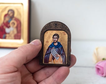 May include: A small wooden icon with a gold background and a painted image of a man in a blue robe holding a scroll. The icon has a dark brown wooden frame with intricate carvings.