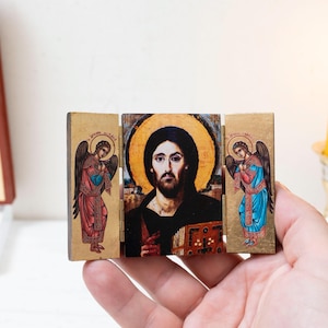 May include: A small, wooden triptych icon featuring a central image of Jesus with a halo, flanked by angels. The artwork is painted in vibrant colors, with gold accents. The icon is held in a hand, with a lit candle in the background.