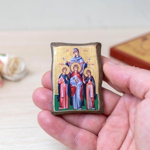 May include: A small, rectangular religious icon featuring a gold background and images of figures in robes. The icon has a dark brown border and is held in a hand. A second icon is visible in the background.