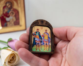 May include: A small wooden framed icon with a gold background depicting two figures in blue and gold armor holding spears. The figures are standing on a green field with a blue sky in the background.