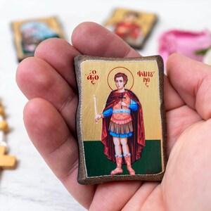 May include: A small, rectangular icon featuring a saint with a halo, holding a candle and a cross. The saint is depicted in a red cape and blue tunic. The icon is held in a hand, with a rosary and other icons visible in the background.