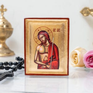 Può includere: Un'icona in legno con sfondo dorato che raffigura Gesù Cristo con una corona di spine e una veste rossa. L'icona è incorniciata in una cornice di legno rossa e ha il testo "IC XC" scritto in lettere dorate.