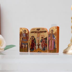 Może przedstawiać: Złoty tryptyk religijny przedstawiający sceny biblijne, w tym ukrzyżowanie, z postaciami w kolorowych szatach. Po prawej stronie zapalona świeca w złotym świeczniku. Białe r&oacute;że i ikona w tle.