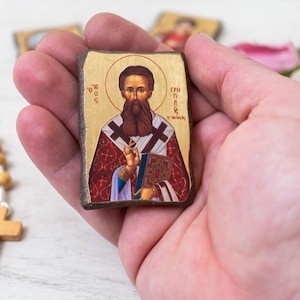 Könnte beinhalten: Eine kleine, rechteckige Ikone mit einer religiösen Figur mit langem Bart, die ein Buch hält. Die Ikone hat einen goldenen Hintergrund und wird in einer Hand gehalten. Ein hölzerner Rosenkranz und ein Kreuz sind im Hintergrund zu sehen.