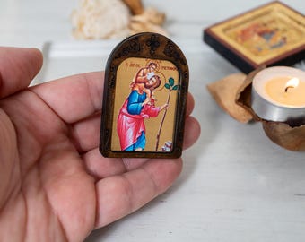 Pode incluir: Um pequeno ícone emoldurado em madeira que representa uma figura religiosa carregando uma criança nos ombros. O ícone tem um fundo dourado e a figura está vestindo uma túnica vermelha. O ícone é segurado em uma mão.