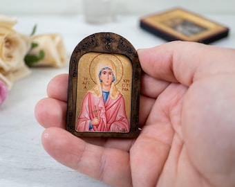 May include: A small wooden icon with a gold background depicting a woman in a pink robe holding a cross. The icon has a dark brown wooden frame with ornate details and the text "Η ΑΓΙΑ ΧΡΙΣΤΙΝΑ" in Greek.