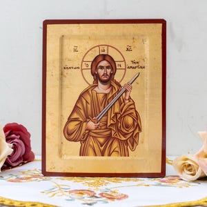 Könnte beinhalten: Religiöse Ikone mit einer Figur mit Heiligenschein, die ein Schwert hält. Die Ikone hat einen goldenen Hintergrund und einen dunkelroten Rahmen. Die Figur ist in goldene Gewänder gekleidet. Das Bild ist von Blumen umgeben.