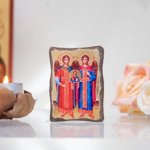 May include: A religious icon featuring two angels flanking a central figure, likely a depiction of a religious scene. The icon has a textured, irregular border. The angels wear red and blue robes. A lit candle and flowers are in the background.