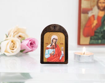 May include: A small wooden icon with a gold background depicting a woman in a red and blue robe holding a cross and a rose. The icon is framed in dark wood and has a rounded top.