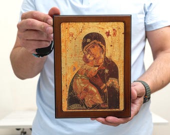 May include: A framed religious icon depicting a mother and child. The icon features warm tones of gold, brown, and red, with intricate details and a dark brown wooden frame. The image is held by a person.
