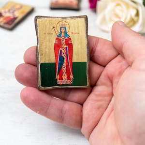 Può includere: Una piccola icona rettangolare tenuta in mano, raffigurante una figura religiosa. L'icona ha uno sfondo dorato, una veste rossa e un mantello blu. La figura è incoronata e tiene una croce. La parte inferiore dell'icona è verde.